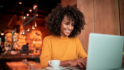Una joven está sentada en una cafetería frente a una computadora. Sonríe al ver que su tienda en línea está en los primeros puestos de los resultados de búsqueda.